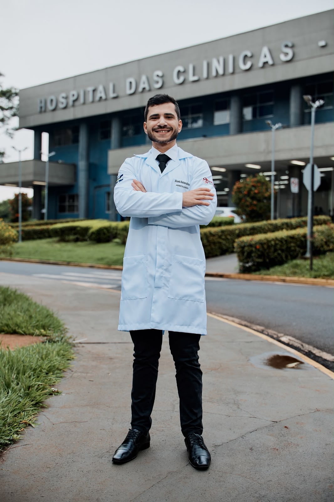 Dr. Ricardo Batista de jaleco, braços cruzados, em frente ao Hospital das Clínicas de Ribeirão Preto.